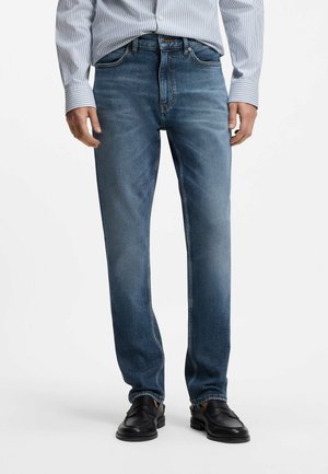 Mand iført blå straight-fit jeans, sorte læder loafers og en lyseblå stribet skjorte med knapper og nedrullede ærmer.