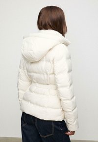 Žena okrenuta leđima nosi bijelu kapu puffer jaknu i tamnoplave traperice pred jednostavnim pozadinom.