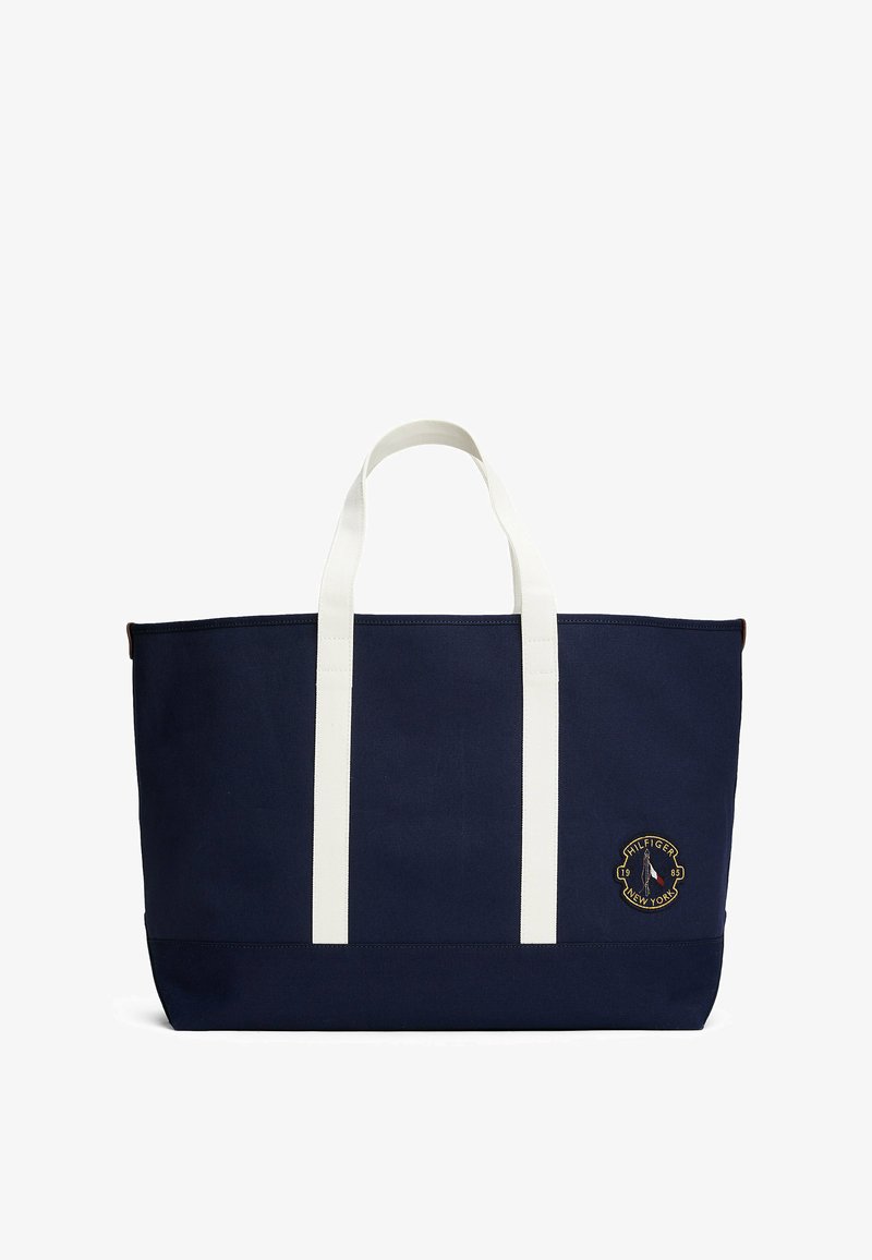 Granatowa torba płócienna z białymi uchwytami, z okrągłym haftowanym logo. Strukturalnie prostokątny kształt z delikatną fakturą.