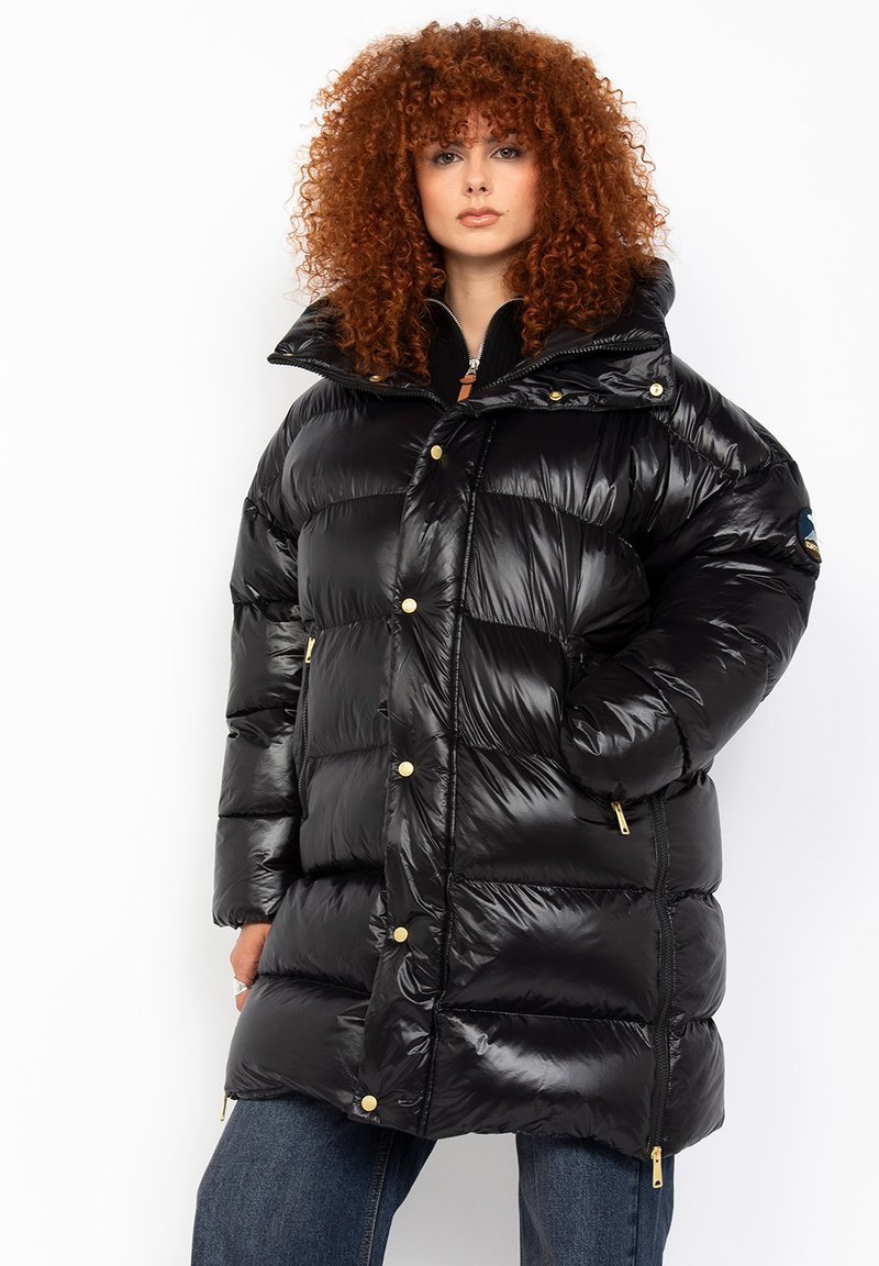Zwarte pufferjas van glanzend materiaal, oversized silhouet, hoge kraag en gouden hardwaredetails. Beschikt over grote, gestikte secties en zakken.