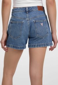 Shorts in denim con orlo sfrangiato, lavaggio blu, vita alta e due tasche posteriori; presenta una patch in pelle con logo e dettaglio del brand.