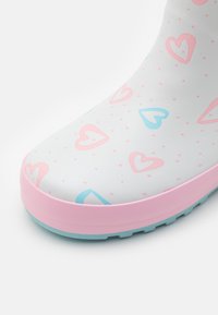 Botte en caoutchouc blanche avec des motifs de cœurs roses et bleus, semelle rose et détails à pois subtils. Bout arrondi et semelle extérieure à texture antidérapante.