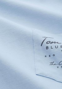 Svetlomodré bavlnené tričko s obdĺžnikovým látkovým štítkom s tmavosivým textom, ktorý obsahuje slová "Tom," "Blue" a kruhový dizajn loga.