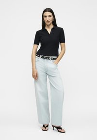 Γυναίκα που στέκεται με το ένα χέρι στην τσέπη ενός ανοιχτού μπλε τζιν με φαρδιά πόδια, φοράει μαύρο κοντομάνικο πουκάμισο με γιακά, ζώνη και μαύρα σανδάλια με τακούνι.