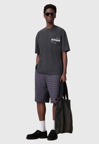 Mørkegrå oversized t-shirt med "ALLSAINTS" grafik, ternede marineblå og brune shorts, sorte ruskindssko, hvide ankelsokker og en sort tote bag.