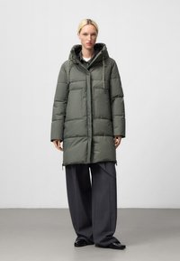 Piumino verde oliva, dotato di cappuccio, due grandi tasche frontali, coulisse regolabile e un taglio dritto e oversize.