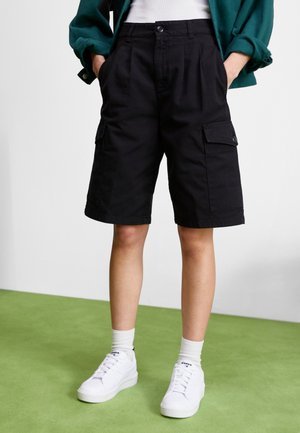 Shorts cargo noirs avec poches latérales, design plissé et fermeture à bouton, associés à des baskets blanches et des chaussettes mi-mollet sur un sol vert.