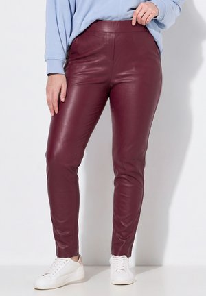 Kvinde iført bordeaux bukser i imiteret læder, lyseblå sweatshirt og hvide sneakers, der står op ad en hvid væg.