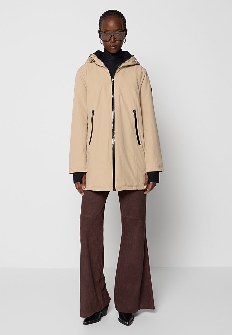 Beige waterdichte jas met een capuchon, voorkant met rits en twee zijzakken. Heeft een ontspannen pasvorm en zwarte accenten.