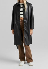 Svart, glänsande konstläderjacka med krage, knappstängning och två framfickor; kombinerad med bruna kordflorsbyxor och vita sneakers.