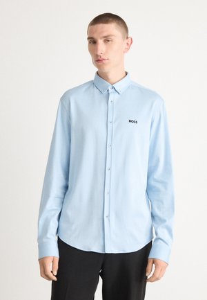 Camisa de manga larga azul claro con cuello abotonado. Presenta un logo negro de "BOSS" en el lado izquierdo y una textura de tela suave.