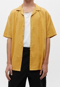 Kurzes gelbes Hemd mit dezentem Muster, knöpfbarem Design und Kragen, getragen über einem weißen T-Shirt und kombiniert mit schwarzen Hosen.