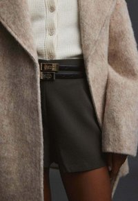 Kabát z béžové vlněné směsi s měkkou texturou, vrstvený přes krémový pletený cardigan a tmavé šortky, s dvěma černými pásky s kovovými přezkami.