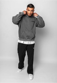 Grå hættetrøje med en frontlomme og sort logo, parret med løse sorte bukser og hvide sneakers, stående mod en ensfarvet baggrund.