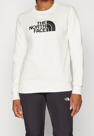 Person trägt weißen Sweatshirt mit langen Ärmeln und schwarze Hose, beide mit schwarzem "The North Face" Logo auf Brust und Oberschenkel.