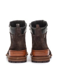 Botas de tobillo de ante marrón con detalles en piel negra, suela resistente y costuras decorativas; presenta un panel de piel con marca en el talón.