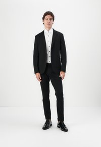 Schwarzer Anzug mit einreihigem Sakko, weißes Hemd mit schwarzen Knöpfen, schlanke Hose und glänzende schwarze Schuhe. Glatte Stofftextur.