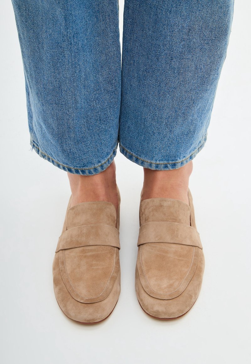 Pieds portant des mocassins en daim beige sans lacets associés à un jean en denim bleu sur un fond blanc uni.