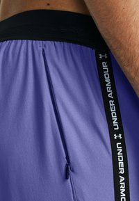Lila Sportshorts mit einem schwarzen Mesh-Bund und einer seitlichen Reißverschlusstasche. Merkmale sind das kontrastierende schwarze "Under Armour"-Branding.