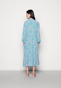 Vestido maxi com um padrão em espiral em rosa e verde sobre um fundo azul-claro. Mangas longas e um corte descontraído, combinado com sandálias de salto bloco rosa.