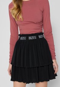 Zwarte geplooide rok met gelaagde textuur en een elastische tailleband met "BIZUU" branding. Gepaard met een nauwsluitend longsleeve top in rozenkleur.