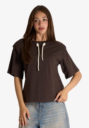 Giovane donna con capelli lunghi che indossa una maglietta marrone a maniche corte con coulisse bianche e jeans denim blu chiaro, in piedi con una mano sui fianchi.