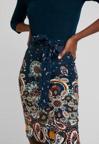 Tygkjol i marinblått mönster med bälte, intrikata blommiga och paisleymönster i livliga färger, med en slät textur och figurnära passform.