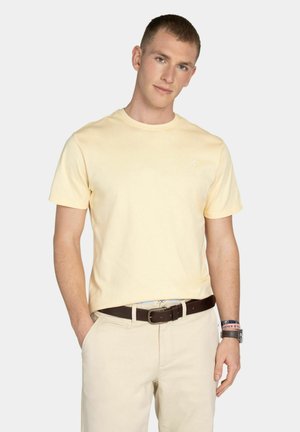 Młody mężczyzna z krótkimi włosami, ubrany w jasnożółtą koszulkę, beżowe spodnie, ciemnobrązowy pasek i akcesoria na nadgarstek, stojący z jedną ręką w kieszeni.