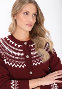 Burgunderfarbener Strickcardigan mit weißem geometrischem Muster, rundem Ausschnitt und silbernen Knöpfen. Weicher Stoff und lockere Passform mit langen Ärmeln.