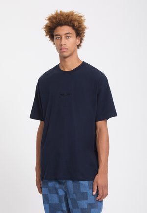 Jeune homme aux cheveux bouclés portant un t-shirt sombre uni et un pantalon à carreaux bleus, debout devant un fond blanc.