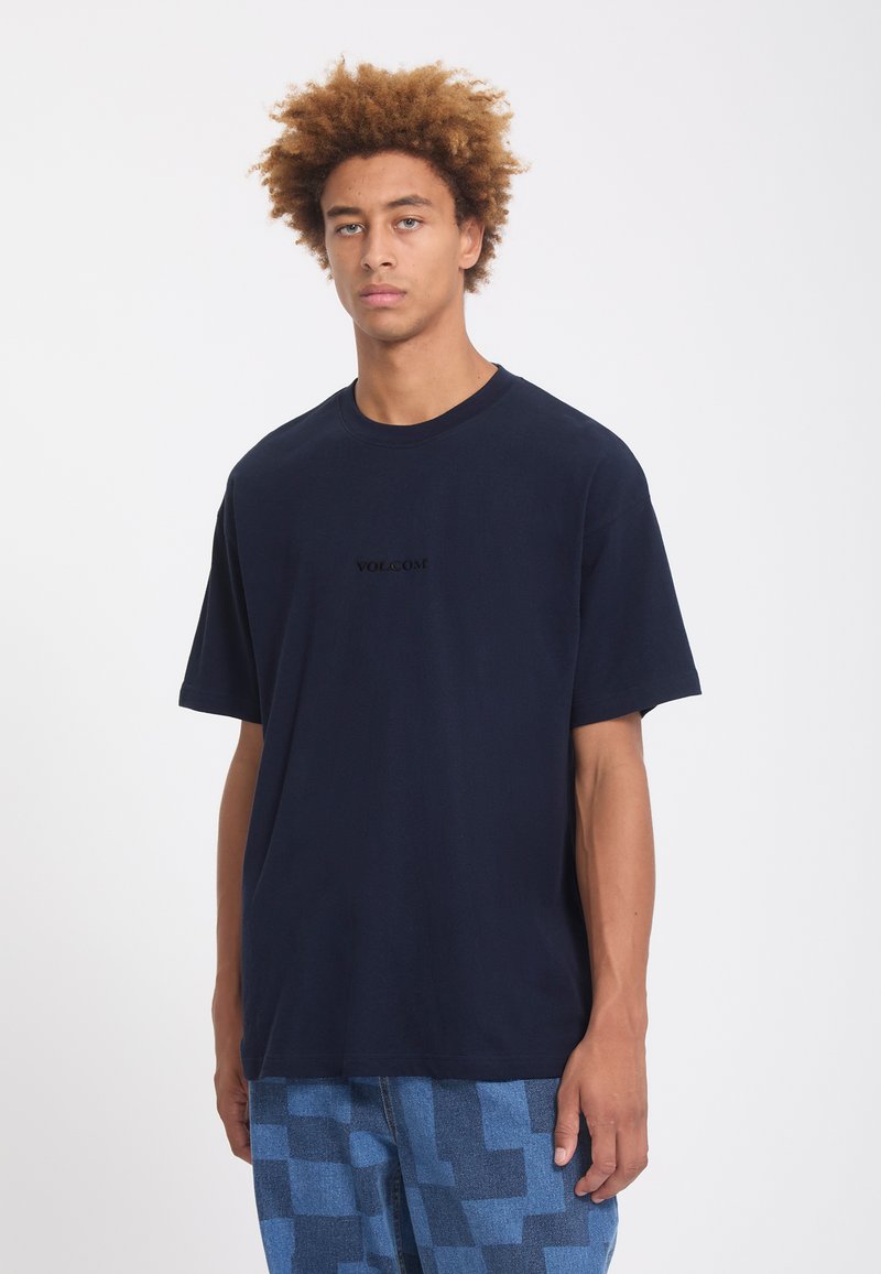 Jeune homme aux cheveux bouclés portant un t-shirt sombre uni et un pantalon à carreaux bleus, debout devant un fond blanc.