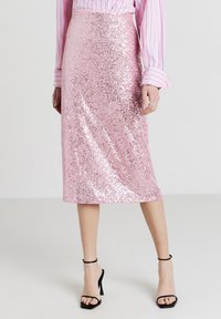 Donna che indossa una gonna midi di paillettes rosa lucide, una blusa a righe rosa e sandali neri con il tacco alto, in piedi su un pavimento chiaro e semplice.
