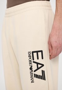 Beige sweatpants made of soft fabric, featuring an elastic waistband and side pocket. Black logo text reads "EA EMPORIO ARMANI" on the leg.