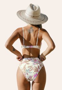 Bikini floral con tirantes ajustables que presenta una base crema y patrones en rosa y azul. La parte inferior de cintura alta ofrece una cobertura completa. Sombrero de paja al fondo.