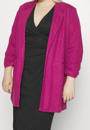 Fuksia ylisuurena bleiserinä, jossa on teksturoitu kangas, lovelliset kaulukset ja irtonaiset kolmannes-hihat, käytetty mustan mekon päällä.