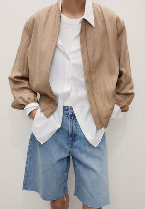 Giacca bomber beige con polsini elasticizzati, indossata sopra una camicia bianca, abbinata a shorts di denim chiaro, con una vestibilità rilassata e lievi effetti di usura.