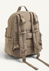 Béžový vankúšový batoh s nastaviteľnými popruhmi, bočnými vreckami a horným uchom. Má hladkú nylonovú textúru a minimalistický dizajn.