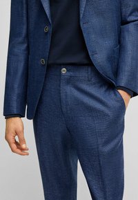 Navy blue textured suit jacket with two buttons, fitted style. Pants match, featuring a single button and side pockets. Fabric appears woven.