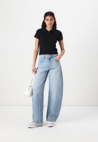 Svart kortärmad polot-shirt, ljusblå vida jeans, vita sneakers och en vit handväska, mot en enkel ljus bakgrund.
