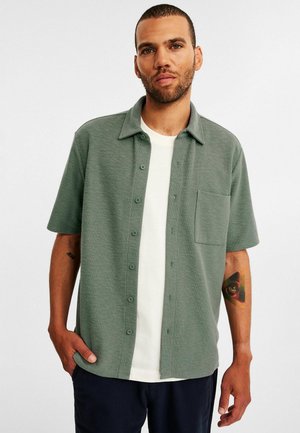 Grøn kortærmet skjorte med knaplukning og lomme, lavet af struktureret stof, parret med en hvid t-shirt under og navy bukser.