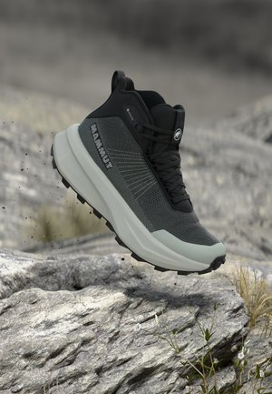 Cizme de drumeție negre și gri cu talpă albă groasă în aer deasupra terenului stâncos, având crampoane rezistente și logo-ul „MAMMUT” pe lateral.