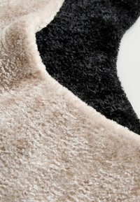 Textured rug featuring a curved design with a soft beige section and a contrasting soft black section, both with a plush finish.