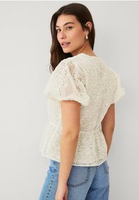 Hvid blonde peplum top med puffede ærmer, indviklede blomster mønstre, en rund halsudskæring og struktureret stof. Båret med lyseblå jeans.