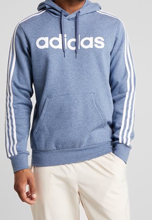 Mann trägt blauen Adidas-Hoodie mit weißen Streifen an den Ärmeln und großer Fronttasche, kombiniert mit beigen Hosen.