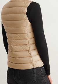Beżowa kamizelka puchowa z poziomym pikowaniem, wykonana z lekkiego materiału. Nosi się ją na czarnej bluzce z długim rękawem, podkreślającej dopasowany kształt.