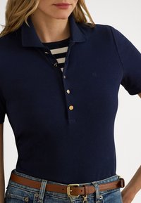 Κοτλέ μπλουζάκι πόλο σε ναυτικό μπλε χρώμα με γιακά, χρυσά κουμπιά και διακριτικό λογότυπο. Φορεμένο πάνω από ριγέ πουκάμισο, συνδυασμένο με τζιν και καφέ ζώνη.