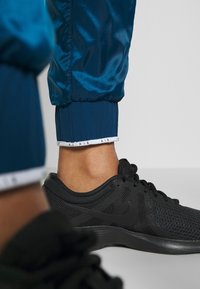 Dunkelblaue sportliche Jogginghosen mit glänzender Textur und schmal zulaufenden Säumen, versehen mit weißem "AIR"-Branding am Saum, kombiniert mit schwarzen texturierten Sneakers.