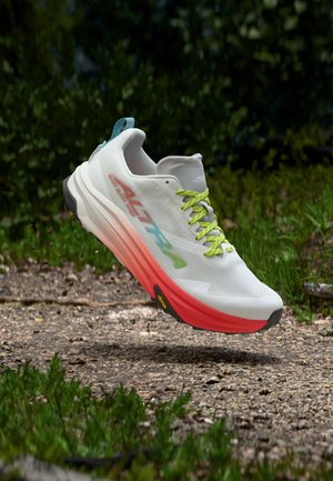 Chaussure de trail running blanche et rouge avec des lacets verts flottant au-dessus d'un chemin en gravier entouré d'herbe et d'arbres.