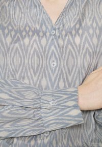 Blusa azzurra chiara con motivo ikat in diverse tonalità, presenta un collo a V, chiusura a bottoni e maniche lunghe con dettagli arricciati.