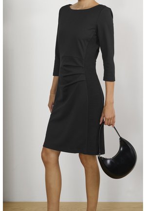 Femme portant une robe noire mi-longue à manches trois-quarts, tenant un sac à main noir en forme de croissant par sa petite anse.